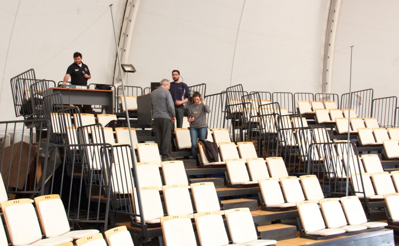 Se observa en el auditorio a cuatro perssonas, probando la colocación e instalción del aro magnético en el auditorio carpa compuesto por gradas y sillas en forma escalonada.